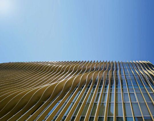 A photo of a very tall building with a glass facade. The building has many windows in vertical rows. The sky is blue and clear.